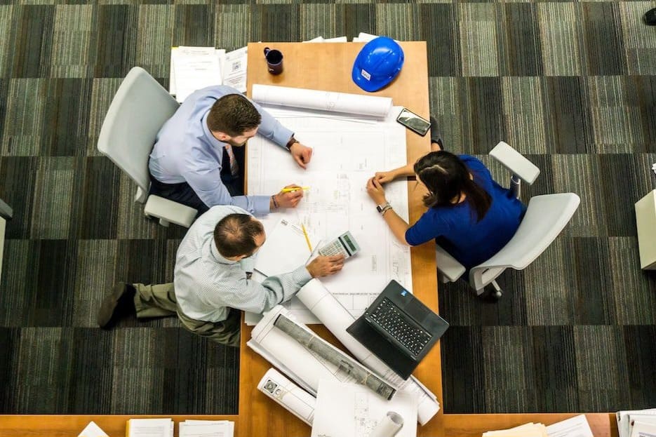 Três profissionais trabalham em um projeto em uma mesa com notebook, papéis, capacete de construção, lápis e calculadora